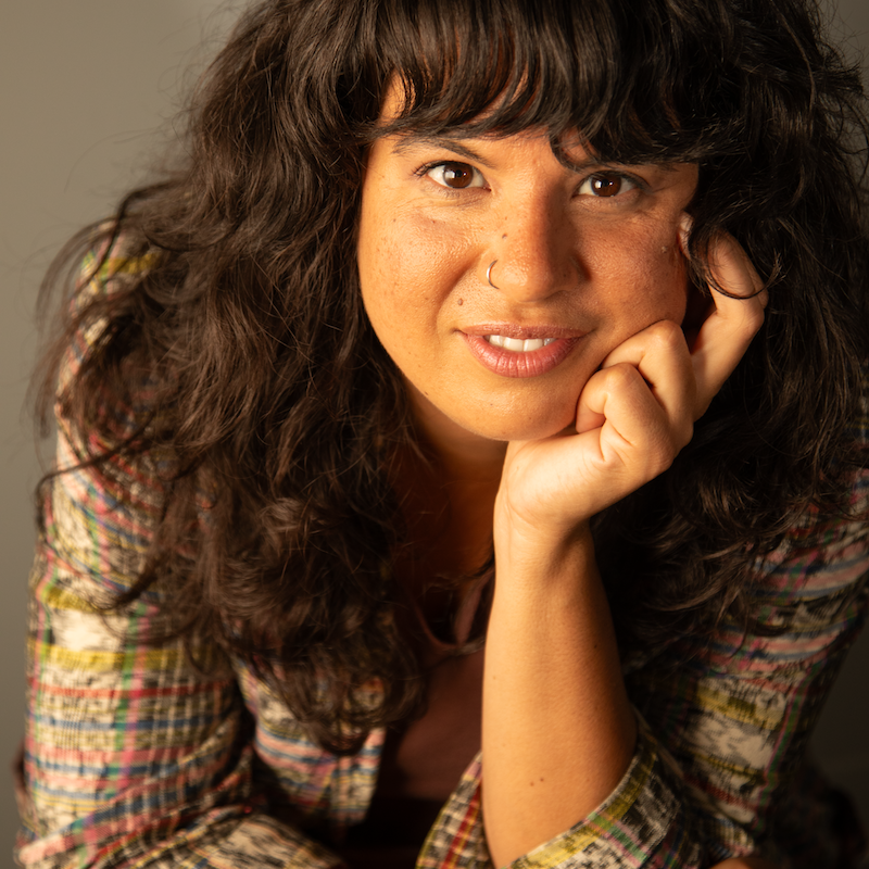 Image shows a person with long dark hair and bangs, with their hand under their chin and smiling