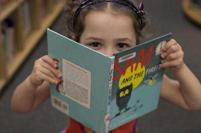 Child holding a book titled "SAM AND THE FIREFLY" in a library.