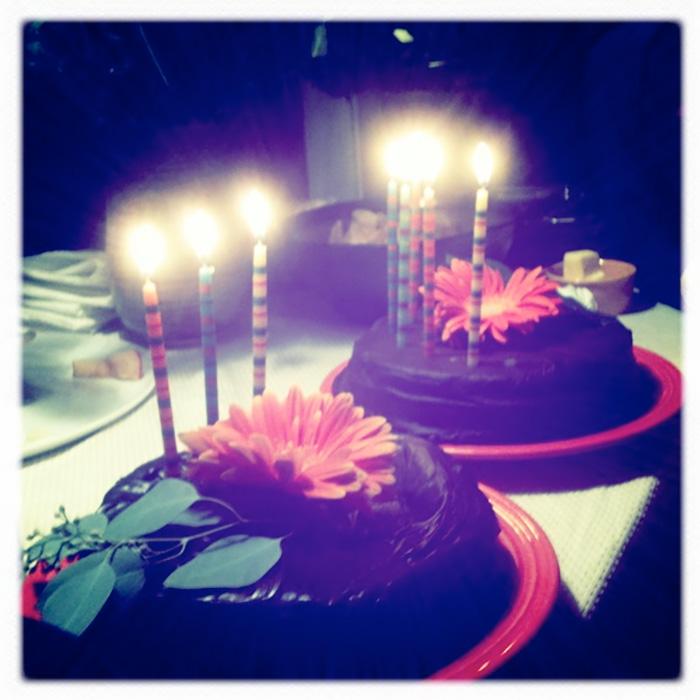 Two chocolate cakes with orange flowers and lit striped candles on red plates.