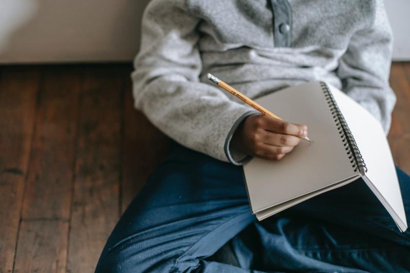 Child sitting cross-legged with a pencil and a blank notebook on a wooden floor.