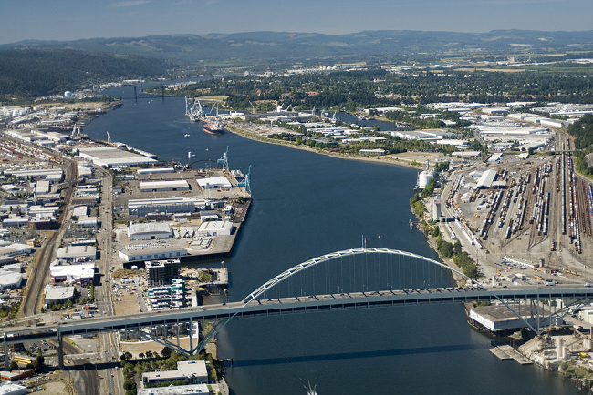 Aerial view of a river with an arch bridge, surrounded by industrial areas and distant green hills.